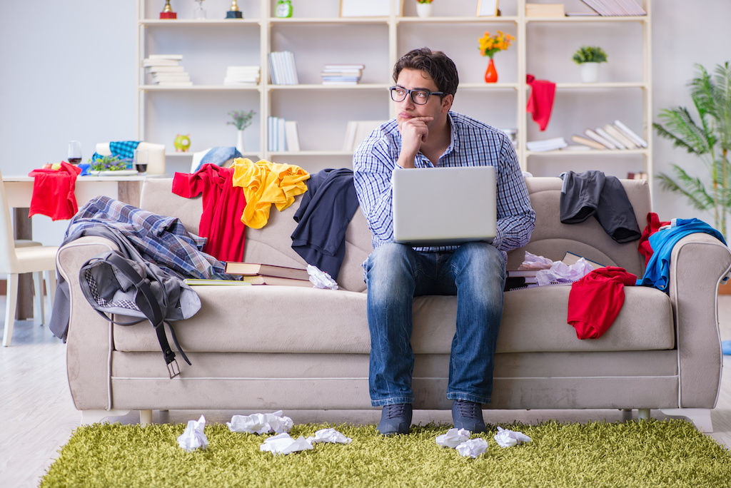 Man sitting on couch with clothes all over the apartment